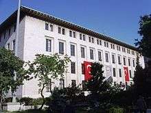 A four-story, white flat-roofed building with two Turkish flags and a portrait on the exterior