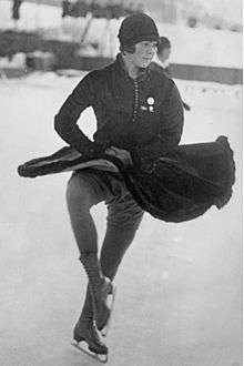 A female figure skater wearing a dark-colored dress and cloche hat performs a turn in an outdoor ice rink.