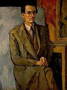 Portrait of artist sitting on chair with partial canvases shown in the background; legs crossed, hands on lap, staring in profile, wearing a blue tie, white shirt, brown vest, brown trench coat, brown slacks, and glasses.