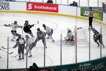 Several hockey players in either white and blue or black and grey uniforms watch as one player shoots the puck into his opponent's net