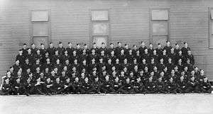 Formal portrait of six rows of men military uniforms, seated or standing in front of a building