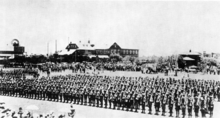 A military unit stands on parade, rifles shouldered, in the middle of a town. Large crowds are gathered around.