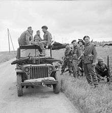 A black and white photograph of a jeep with soldiers sitting on top and standing beside it. The soldiers sitting on the jeep are three German soldiers and one British soldier who is interrogating the Germans. On the bonnet of the jeep is small motorcycle, while in the background is a Horsa glider