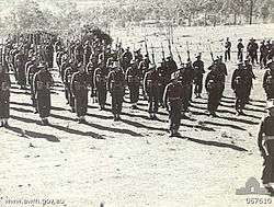 A black and white photograph of soldiers on a unsealed parade ground in a rural setting, wearing battledress uniforms, with slouch hats, webbing and rifles.