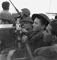 Black and white photo showing men in military uniforms wearing helmets in a very crowded vehicle. The men are pushed up against a large machine gun, and another machine gun is visible at the front of the vehicle pointing at the sky.