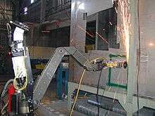 A robot with a metal arm is using its grinding wheel to cut into a wall, sending a shower of sparks upward.