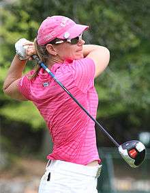 A blonde-haired woman in a pink shirt and hat with white pants and glove and a driver in her hand in the position at the end of a golf swing