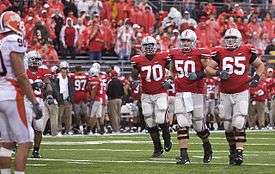 Three football players in red and grey uniforms approach the line of scrimmage.