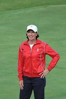 A brown-haired woman in a red jacket and navy blue pants and white undershirt and white hat