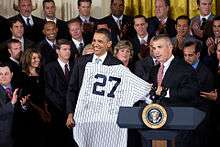 Two men in dark suits stand in front of a crowd in the background and behind a podium in the foreground with the Seal of the President of the United States on it. The two men are holding a white jersey with pinstripes and the number 27 on it.