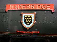 A 'West Country' class enamelled metal nameplate and shield mounted on flat metal casing covering the locomotive boiler. The nameplate comprises a scroll, and below this is a shield containing a picture of a coat-of-arms. A second scroll is below the shield, allowing identification as a member of the 'West Country class'.