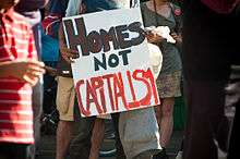 Photograph of a protestor holding a sign saying, "HOMES NOT CAPITALISM"