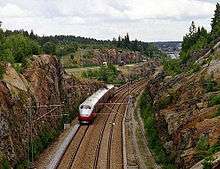 A four-car multiple unit running along a double-track line between two cliffs; in the background is a motorway.