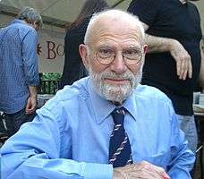 A grey-haired Oliver Sacks with glasses, a beard and a blue shirt with three people in the background
