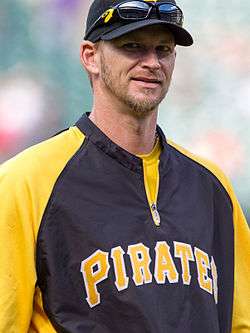A smiling man wearing a black baseball cap with sunglasses attached and a black and yellow baseball jersey with "PIRATES".