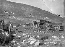 Cart wheels, wagons destroyed barrels and other wreckage in foreground, two soldiers on the road in the middle distance near a staff car