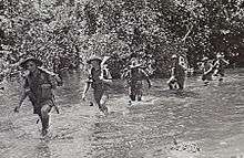 Soldiers wearing slouch hats and shorts wade along a watercourse