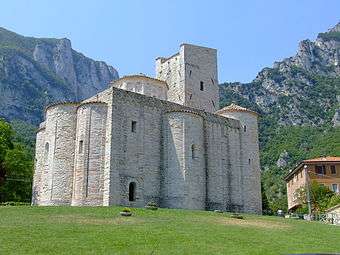 A small church sits on a steep rise, surrounded by craggy mountains. It is basically square with three bulging projections and a castle-like tower.