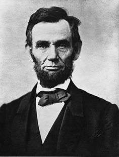 A dark-haired man with a beard and mustache wearing a black coat, white shirt, and black tie