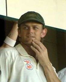 Adam Gilchrist, in white cricket uniform and baggy green hat, holds his left hand to his chin