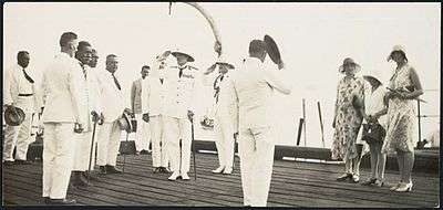 Several men dressed in white tropical uniforms on the deck of a ship, two men wearing pith helmets are saluting, and a group of women wearing dresses and hats are standing to one side