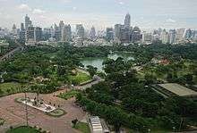 A park with many trees and a lake; a bronze standing statue in front of the park; many buildings in the background