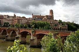 A distant view of a small city in the background and a narrow bridge hidden by a few trees along a river.