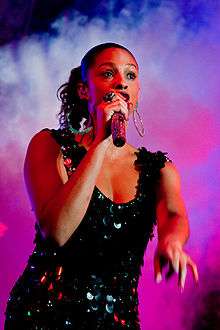 A dark-haired young woman wearing a black dress, singing into a microphone