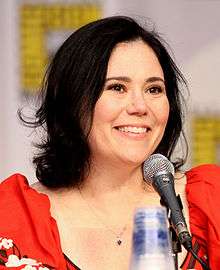 A woman with black hair tied back and light skin, laughing into a microphone, with three vague symbols behind her.