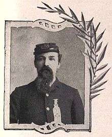 Head and shoulders of a white man with a long beard, wearing a forage cap and a plain jacket with a star-shaped medal and a long ribbon pinned to the left breast.