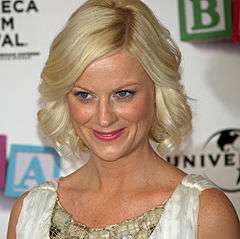 A shot from the shoulders up of a blond woman with blue eyes wearing a white and green dress, smiling and looking at something outside the image