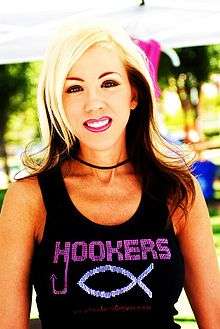 A photograph of a woman standing outdoors while wearing pink lipstick, a black necklace, and a black sleeveless shirt with the word "HOOKERS" in pink and an ichthys in silver