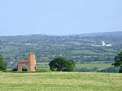 Patchwork of fields and trees with buildings showing in the distance. In the foreground is grass with a ruined building.