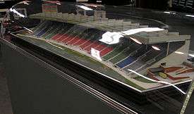 A scale model of stadium seating enclosed within a glass or plastic bubble which reflects an overhead light. There are nine columns of seats in the centre coloured red, two columns on each side of those coloured green, then one column on each side is blue, and one column on each side is grey. The seating is covered by an overhanging roof, and the structure has a concave arc shape.