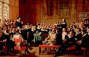 A painting of the Westminster Assembly in session. Philip Nye is standing and gesturing. Various figures are seated around a table. Prolocutor William Twisse is seated on a raised platform.