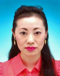 A portrait of an Asian woman with long hair, arranged in front of body on one side, wearing a pink shirt and yellow-orange earrings, looking straight at the camera.