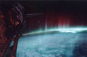 The lower half is the blue-white planet in low illumination. Nebulous red streamers climb upward from the limb of the disk toward the black sky. The Space Shuttle is visible along the left edge.
