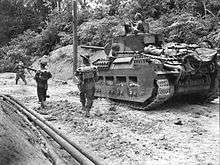 Black and white photo of a tank on a muddy road through tropical terrain with three soldiers walking to the left of it. The upper body of a man wearing a helmet is visible in the turret of the tank.