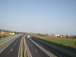 Six lane motorway running through rural area, separated by noise barriers.