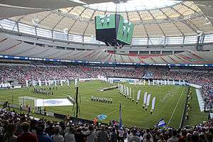 Row of people with white flags stand on a soccer field inside of a stadium. People watch from their seats, which surround the field.