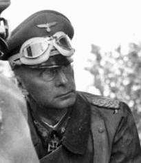 male in German field uniform wearing a peaked cap with goggles resting above the peak
