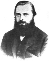 A man in his late 20s or early 30s with dark hair and a bushy beard, wearing a dark coat, dress shirt and tie