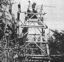 A black and white photograph of a wooden tower in the woods. A man is standing on its open deck