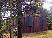 A brown wooden building in a small grasy clearing partially obscured by evergreen trees in the left foreground. It has green bars on the windows and a small pile of wooden debris propped against the front left corner.