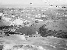 Black and white photograph of a group of aircraft flying in formation above a fjord surrounded by snow-covered mountains