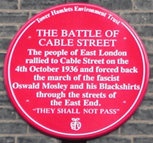 Red plaque from Tower Hamlets Environment Trust reading The Battle of Cable Street. The people of East London rallied to Cable Street on the 4th October 1936 and forced back the march of the fascist Oswald Mosley and his Blackshirts through the streets of the East End. 'They Shall Not Pass'
