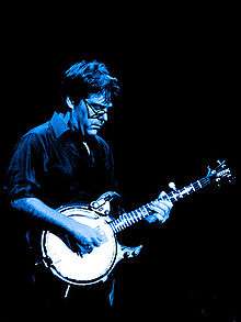 A man wearing a dress shirt and glasses, playing a banjo. A light is shining down on him from above, casting a blue shade over him.