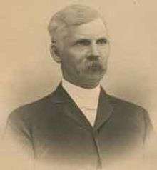 A man in his forties, facing right, with graying hair and a mustache. He is wearing a white shirt and black jacket