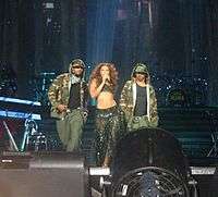 Two men and a woman are looking forward. Both men are wearing military-style clothing, while the woman is holding a microphone. She wears gypsy-style clothing. In the background, many musical instruments are visible.