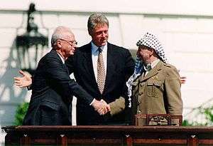 A stolid balding man in a dark suit on the left shakes the hand of a smiling man in traditional Arab headdress on the right. A taller, younger man stands with open arms in the center behind them.